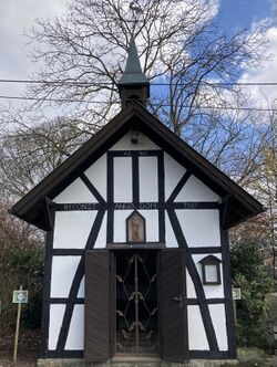 Das Foto vom 16.3.2025 zeigt die Antoniuskapelle in Wahlfeld, die eine Fachwerkkapelle ist mit einem Dachreiter aus Holz, der auch ein kleiner Glockenturm ist. Die Flügeltür der Kapelle ist geöffnet und der Blick fällt auf einen floral verzierten Eintrittsschutz aus Metall, der den Besuchern den Blick in die kleine Kapelle ermöglicht. Im Hintergrund sind unbelaubte Bäume zu sehen, der Himmel ist wolkenreich und bewegt, rechts im Bild ist auch blauer Himmel zu sehen.