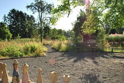 Zu sehen ist links vorne angeschnitten die großen Figuren eines Standschachspiels auf runden Platz, der mit Kies belegt ist, wo Sitzbänke stehen. Im Hintergrund wieder Gartenbeete mit inklusiven Wegen.