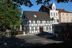 Zu sehen ist das Fachwerkgebäude Alter Zoll mit Aussenterasse der Gastronomie auf der linken Bildseite, ganz links steht ein großer belaubter Baum. Links daneben sind weitere ältere Häuser zu sehen. Im Vordergrund die Straßenkreuzung.